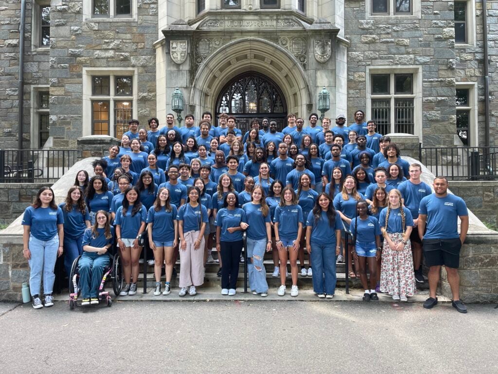 A group of students are standing centered on top of stairs leading into a gray, brick building known as Copley Hall